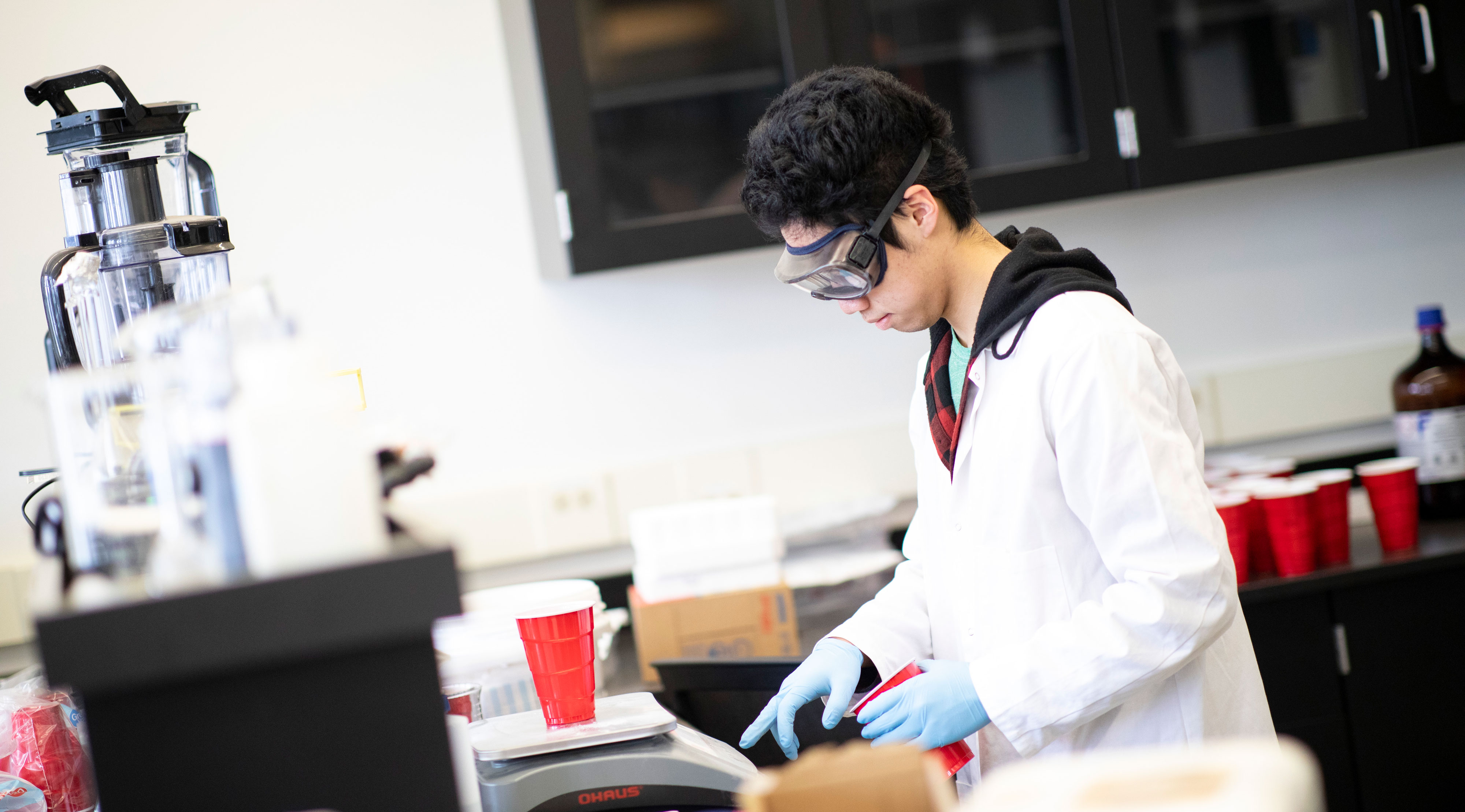 A student wearing safety googles, blue gloves and a white lab coat weighs the contents of a red cup inside a lab.