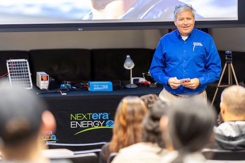 A man in a blue shirt stands in front of an auditorium of UAlbany students next to a table that says "NextEra Energy"