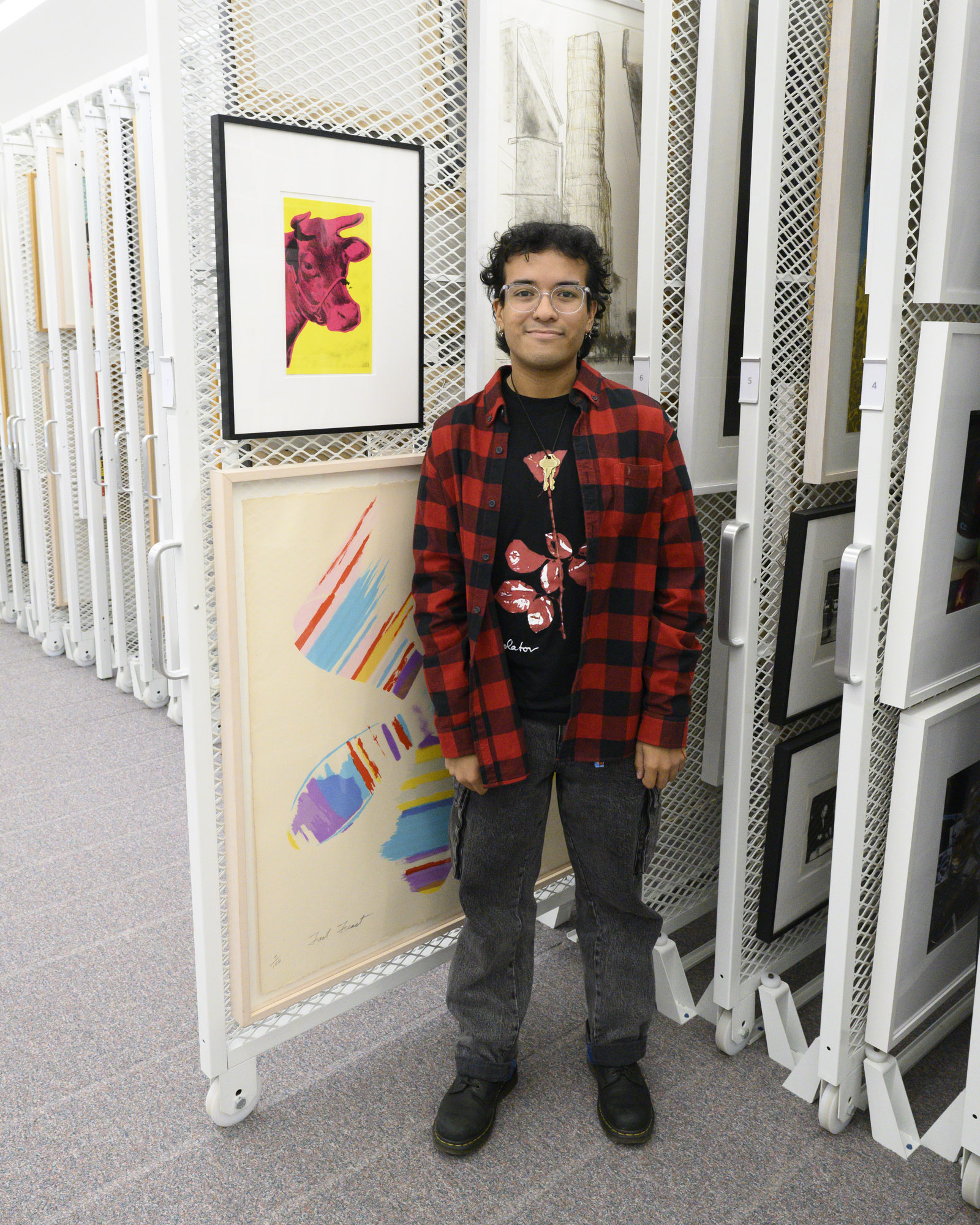 Collections Intern Diego Herrera stands in front of an Andy Warhol screenprint of a fuschia cow head against a bright yellow background. The work is hung on a sliding rack in the Collections Study Space. 