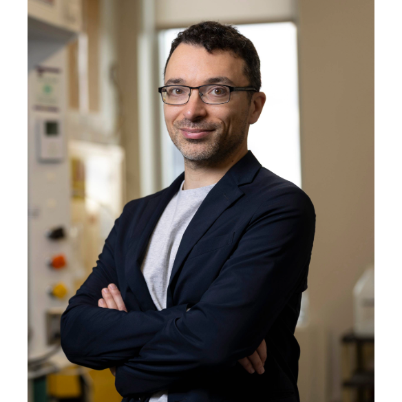 Portrait of a man with short dark hair and glasses, wearing a black blazer.