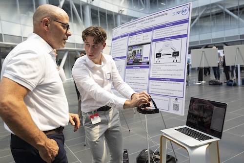A young man with short blonde hair holds a headset and gestures in front of a laptop as an older man looks on.
