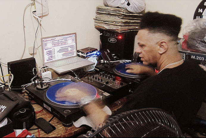 DJ Strike One is seated at a table and performing a scratching routine.