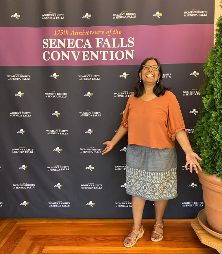 A woman with dark hair and glasses, wearing an orange blouse and gray skirt stands in front of a backdrop that reads, "175th Anniversary of the Seneca Falls Convention," with repeating NYS logos.