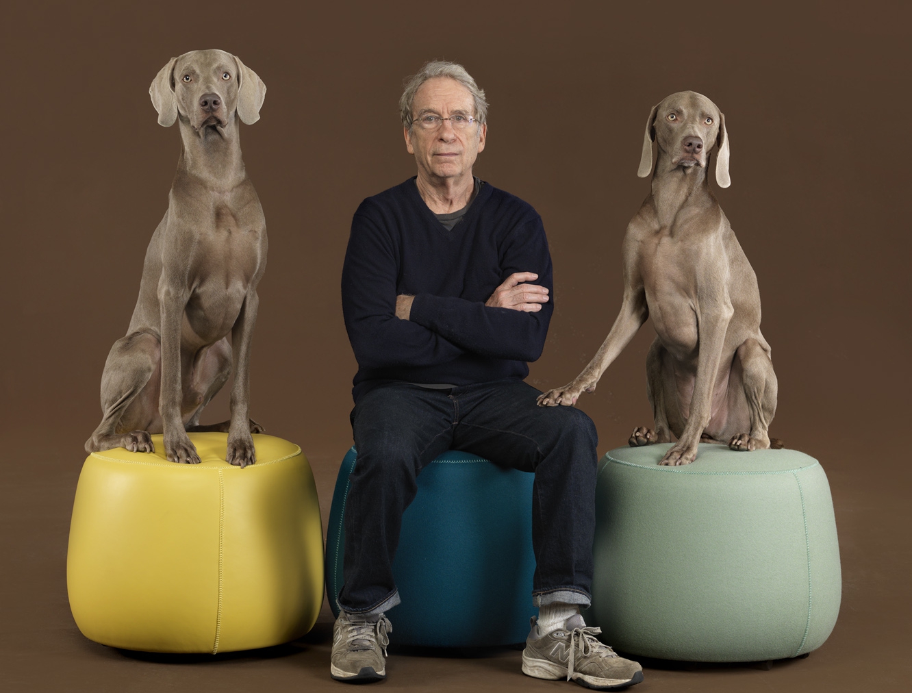 man sits on cushioned stool with arms crossed between two dogs also on stools