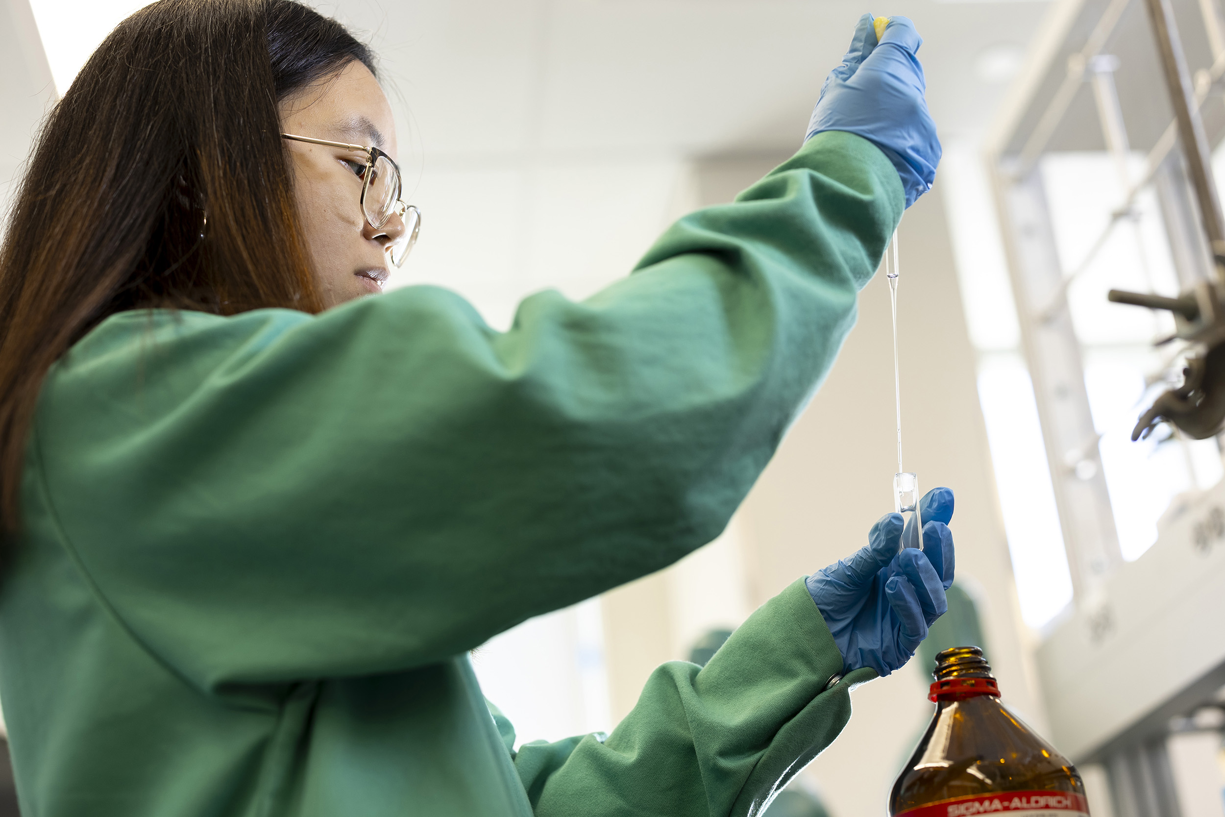 A student with long dark hair and glasses wearing a green lab coat uses a pipette to siphon a clear liquid from a small jar.