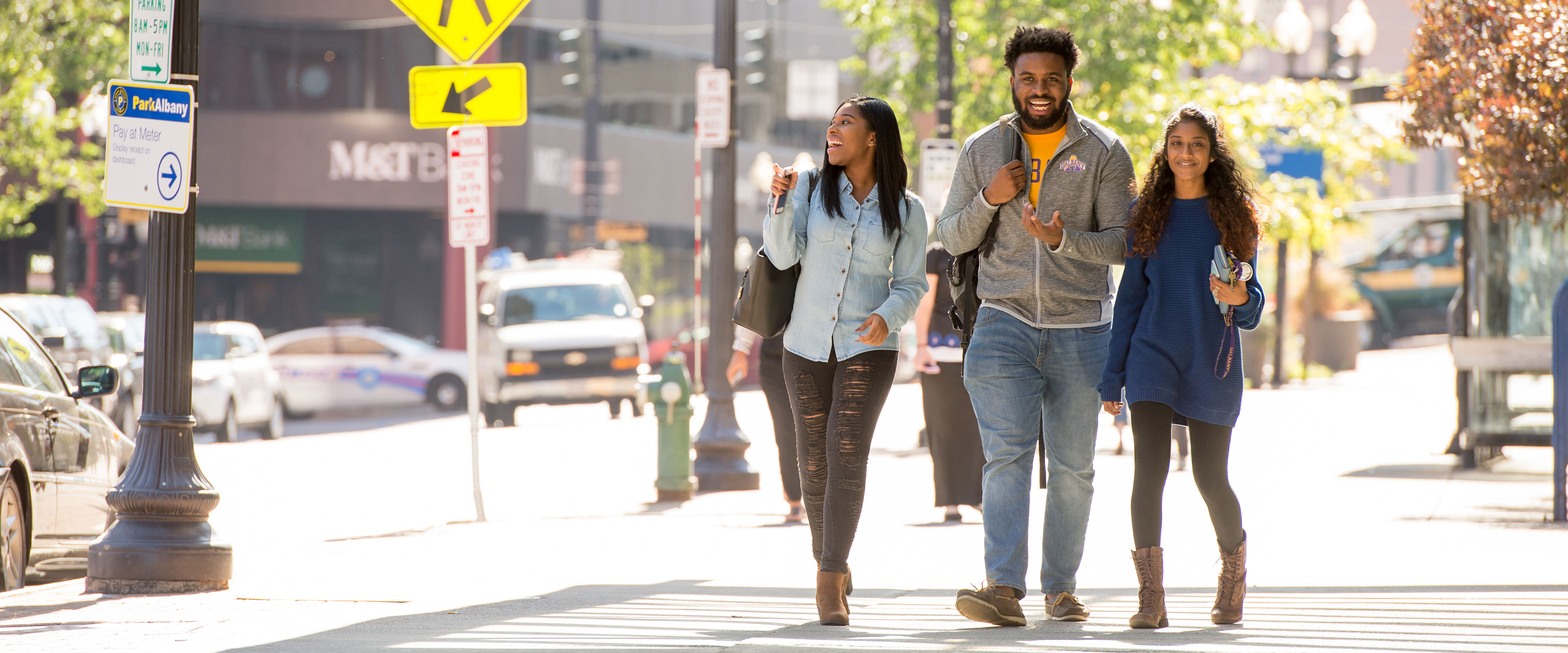 Three UAlbany students smile and laugh as they walk through downtown Albany on a sunny day.