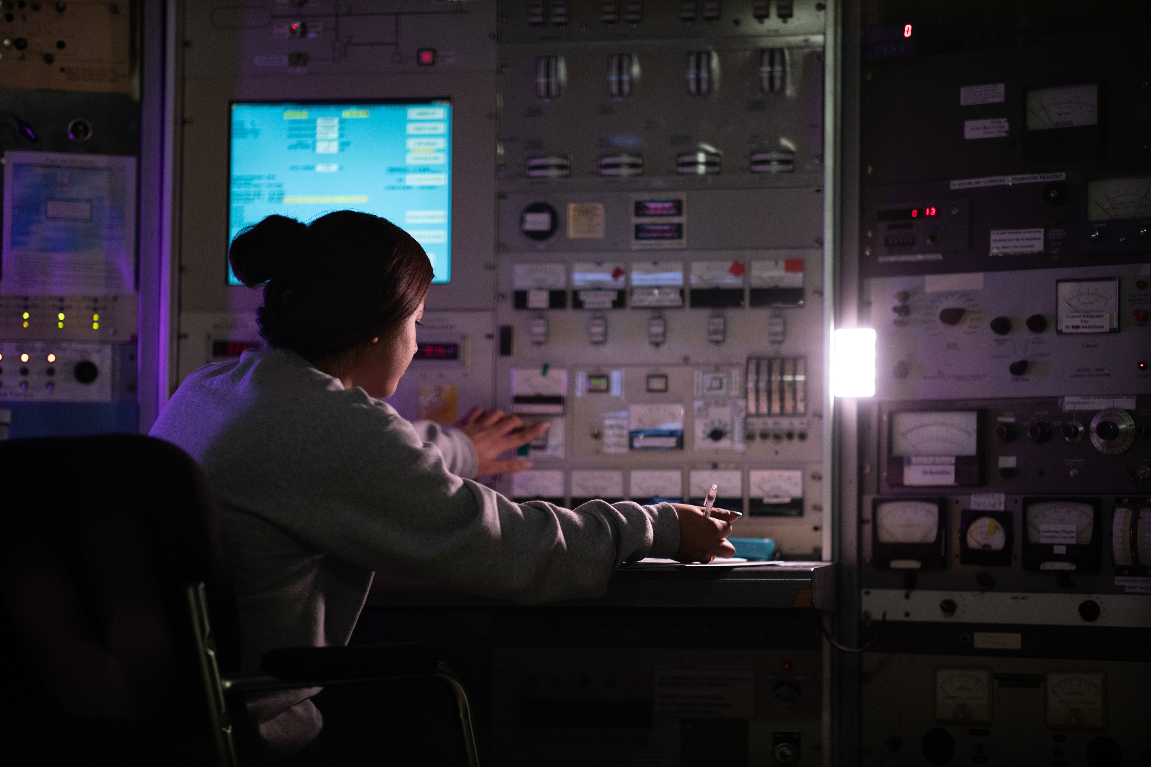 A woman with dark hair in a bun sits in a chair in front of a large instrument panel. With one hand she presses one of the buttons and with the other hand she takes notes.
