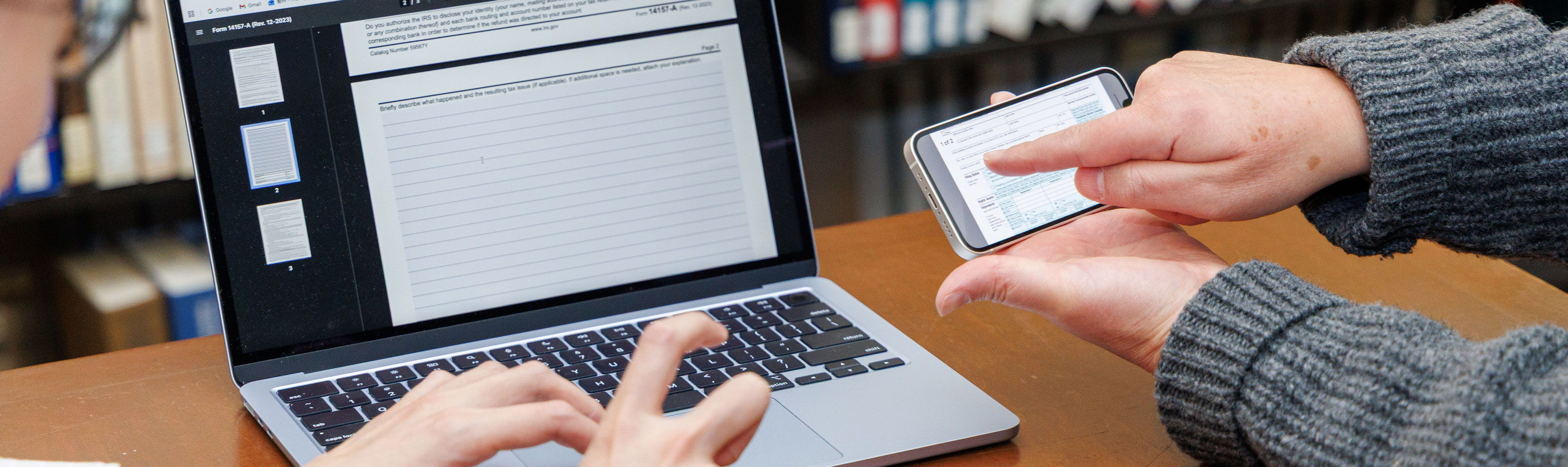 One person types on a laptop while a second person points to something on a smartphone's screen.