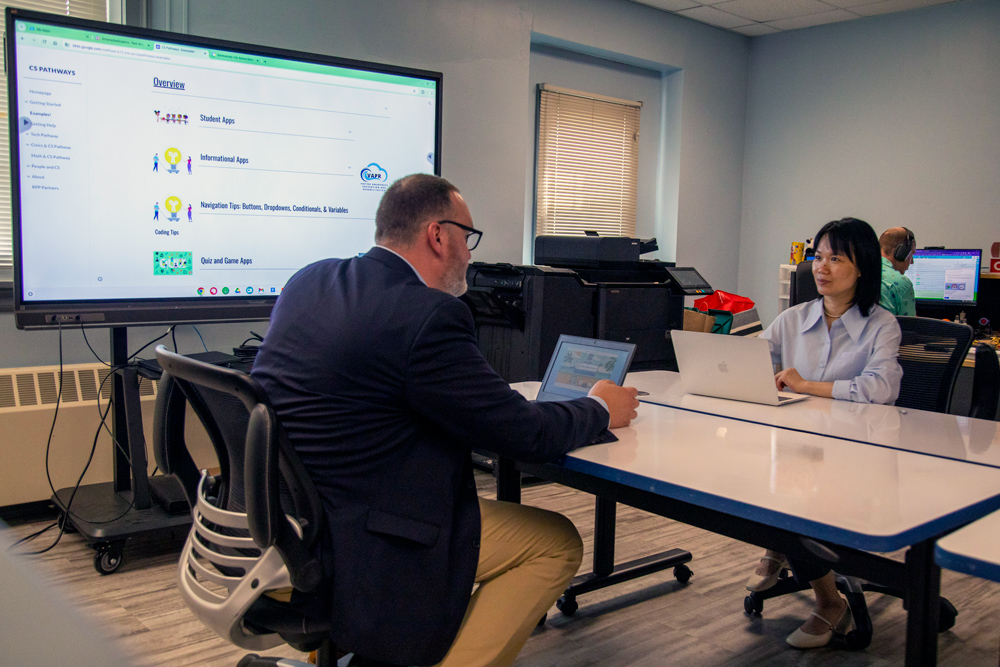 Two educators sitting at a table with laptops and a projection screen.