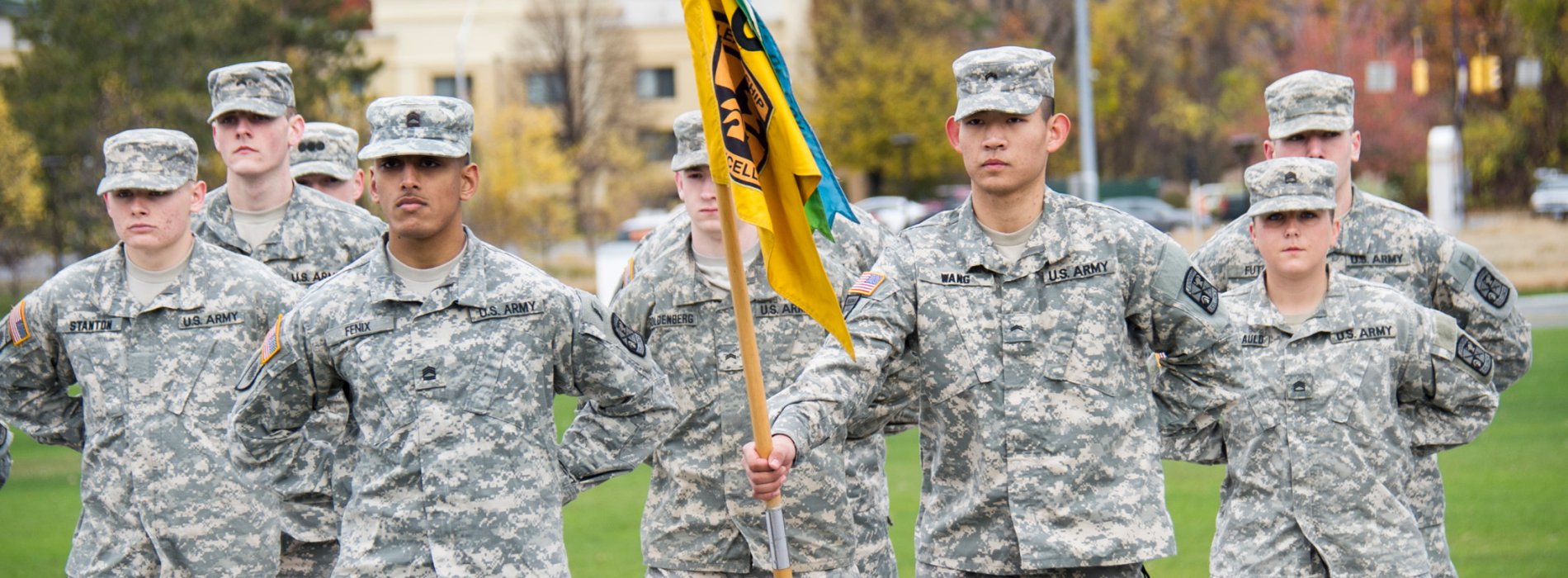 A group of students wearing ROTC fatigues stand in formation. One student is holding a yellow flag.