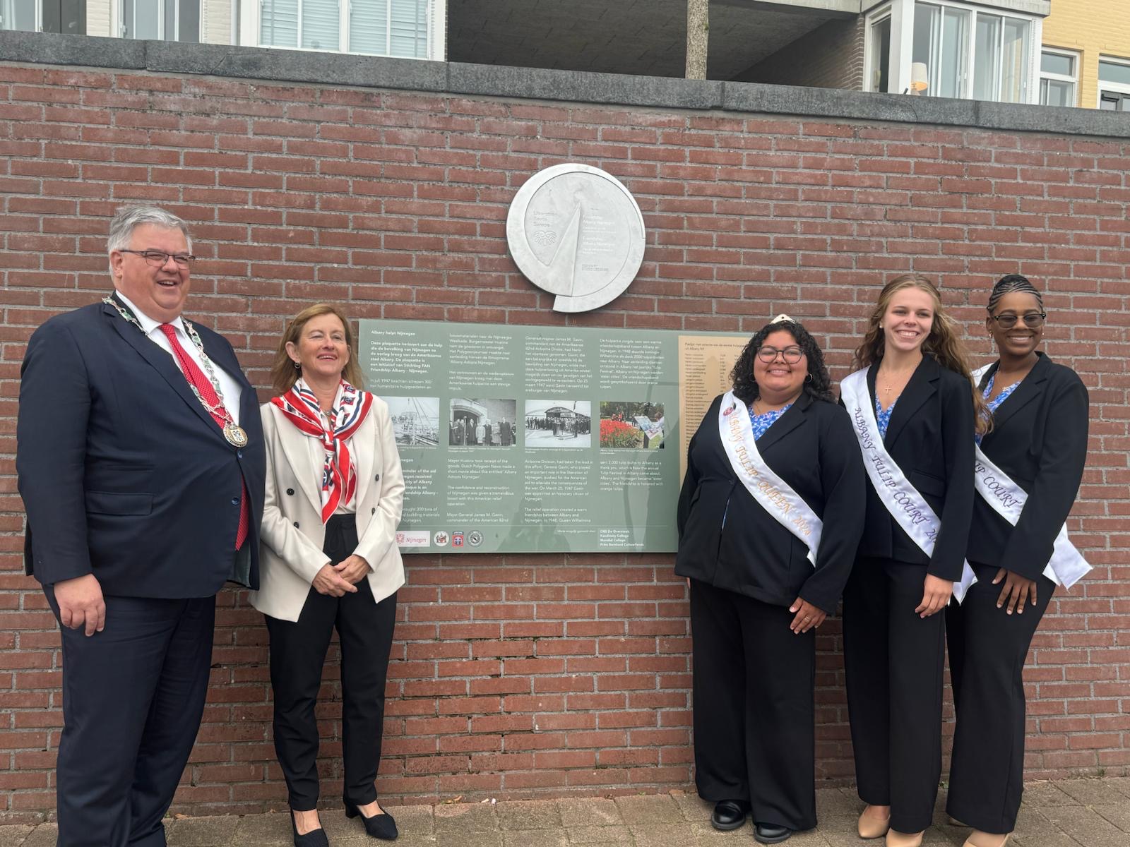 Five people, one man and four women, wearing business attire stand smiling in front of a brick wall bearing an informational panel and large metal seal. 