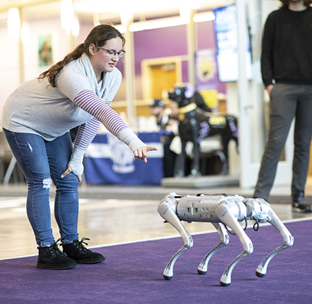 A girl holds her finger out in front of a robot dog inside on a purple carpet.