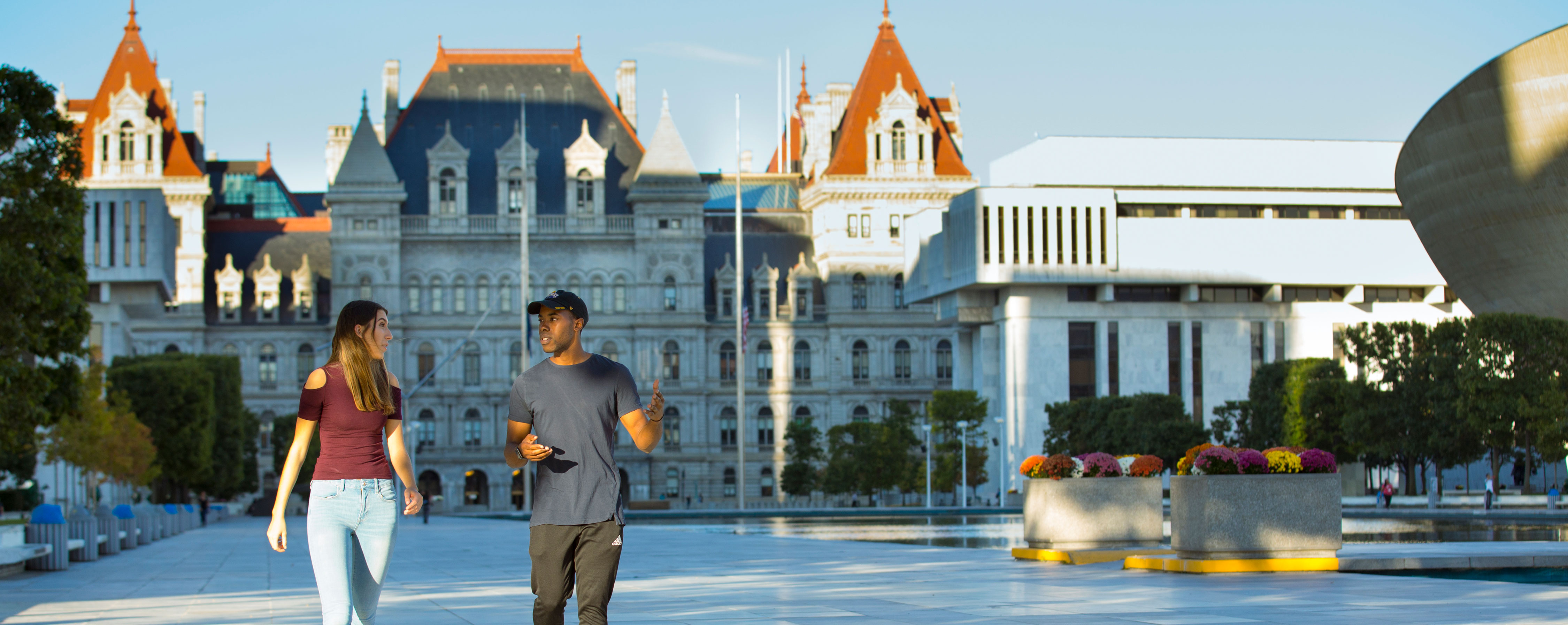 Two students gesture as they talk and walk along the Empire State Plaza, with the New York State Capitol Building behind them.