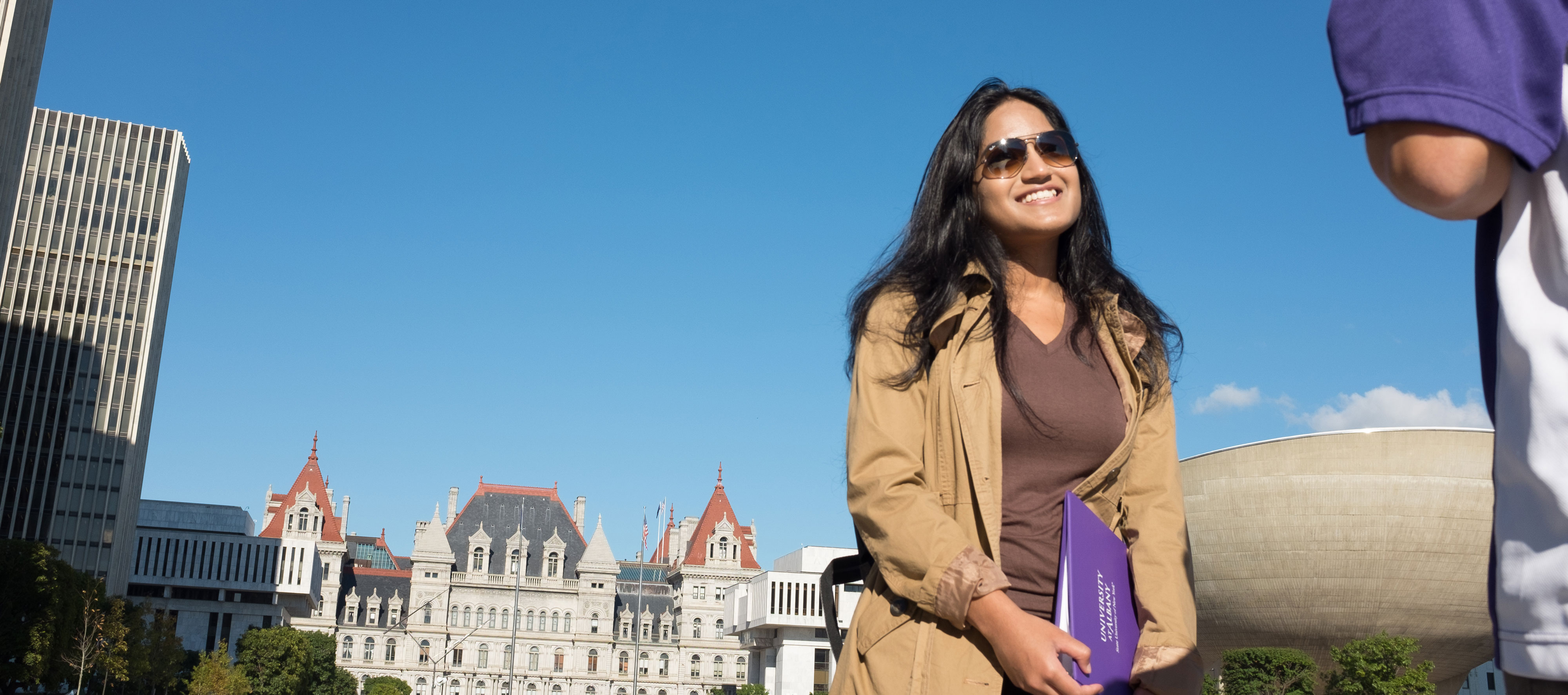 A student wearing sunglasses and a light coat smiles as she stands in front of the New York State Capitol building.