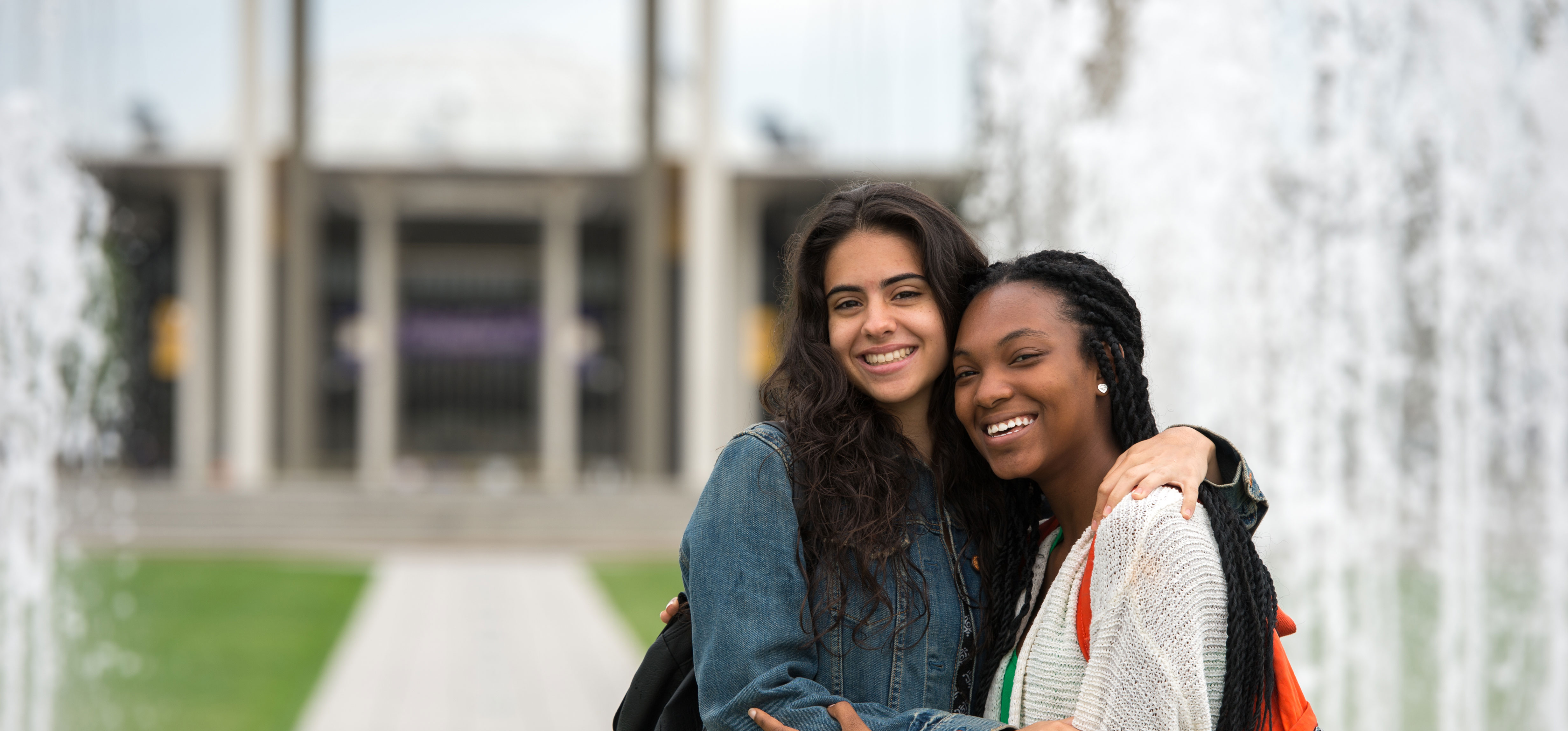 Two students with backpacks embrace as they smile and pose for a photo in front of fountains on the Uptown Campus.