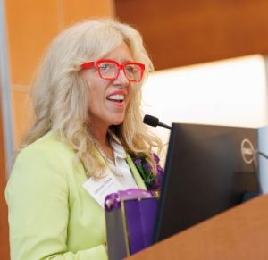 A woman in bright red glasses speaks from a lectern