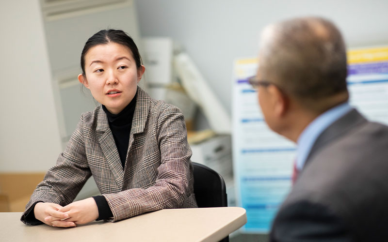 A woman in a black shirt, a black patterned blazer and her hair pulled back sits at a table and speaks with a man in a suit, gray hair and glasses.