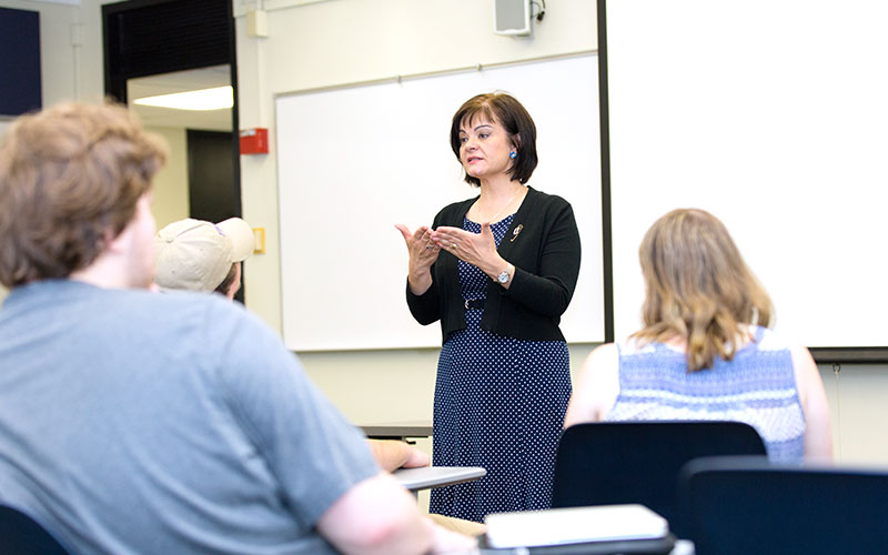 A woman with dark hair, wearing a dark patterned dress and a black cardigan addresses a classroom full of people. Behind her is an empty whiteboard and blank projection screen.