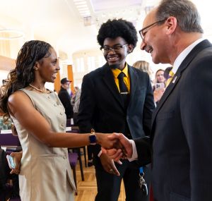 A man in a suit and glasses shakes the hand of a smiling young woman in a pale dress as a young man in a suit and tie smiles at her.