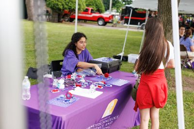 A woman at a table loaded with parts of circuit boards speaks to a girl at an outdoor event