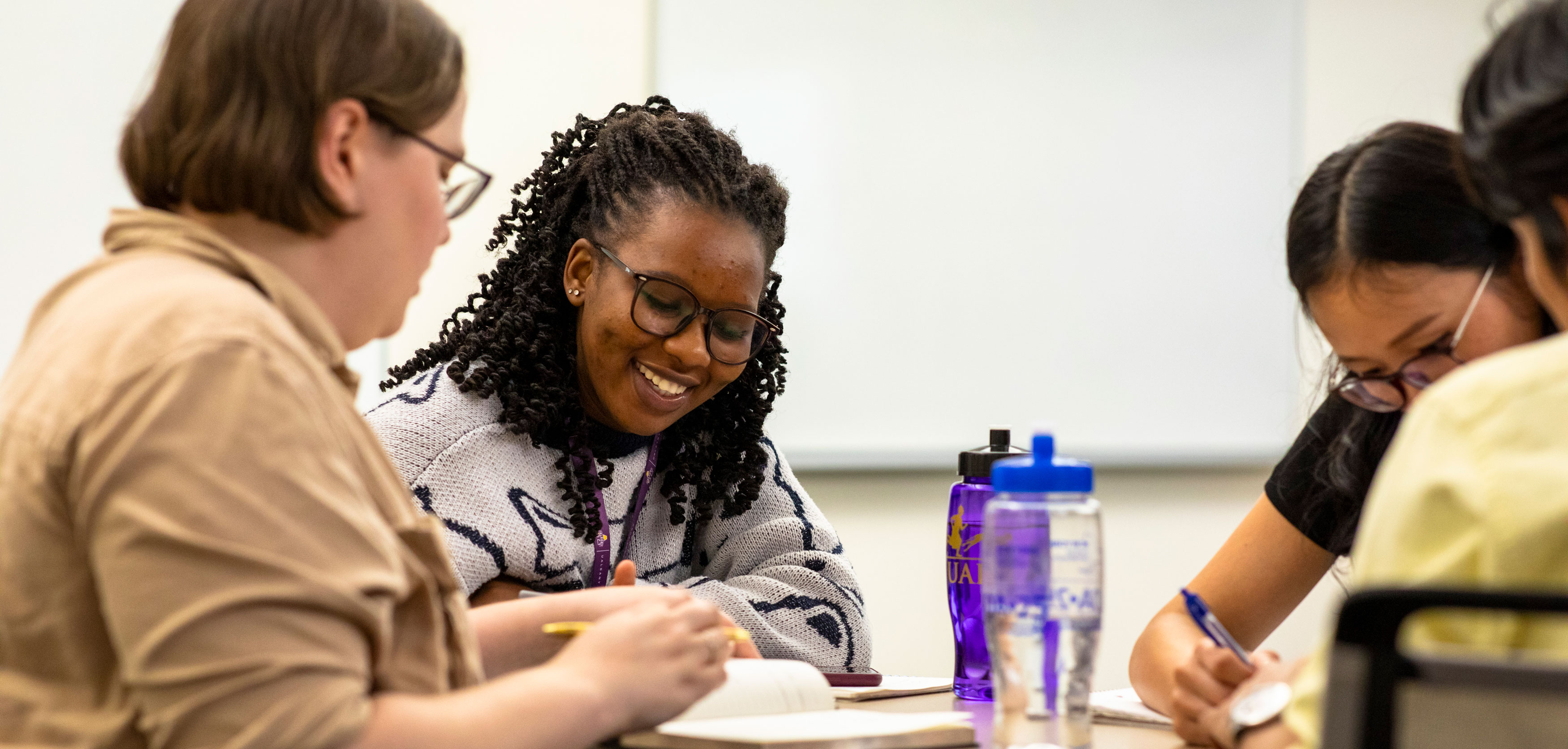 Four students smile as they sit at a table and write in their notebooks together.