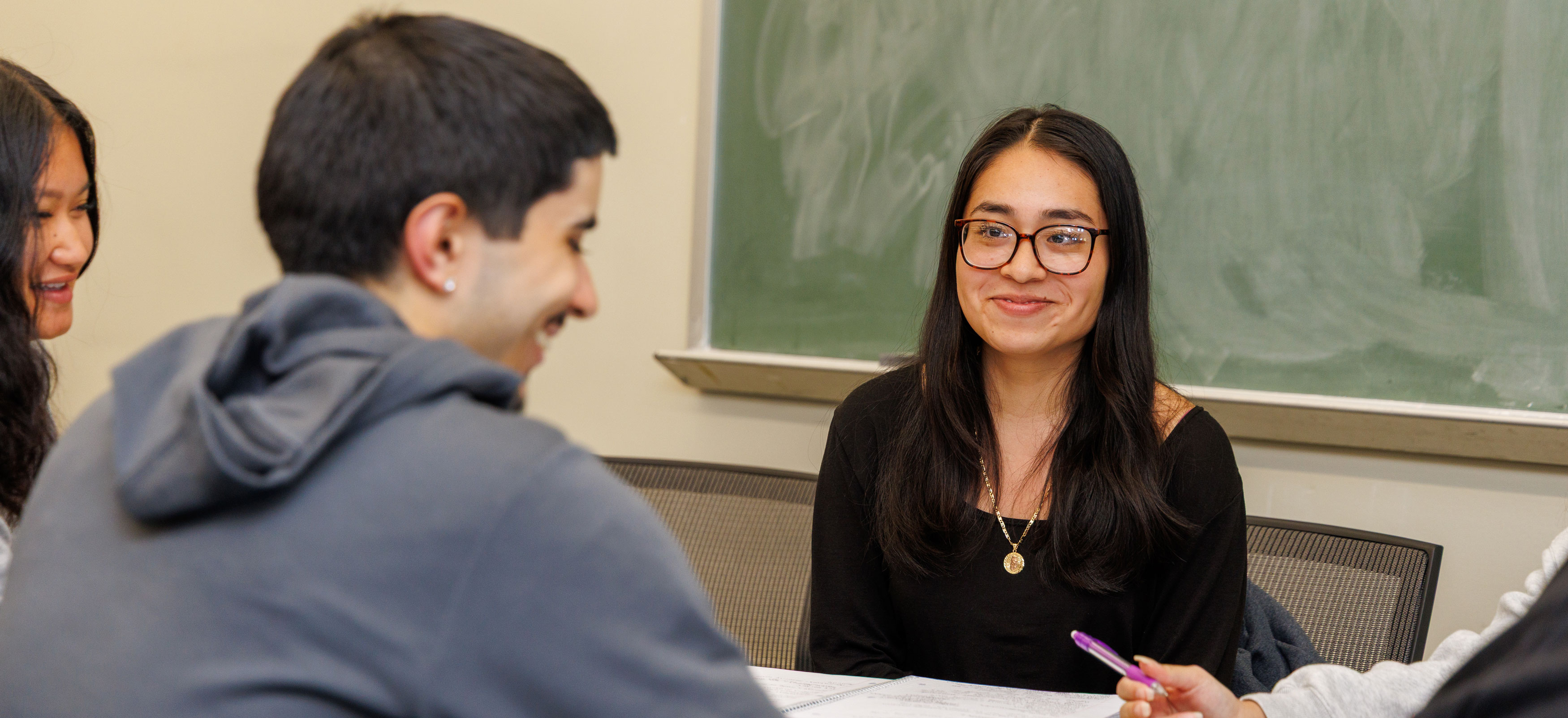 A student seated in front of a chalkboard smiles at another student during a class discussion.