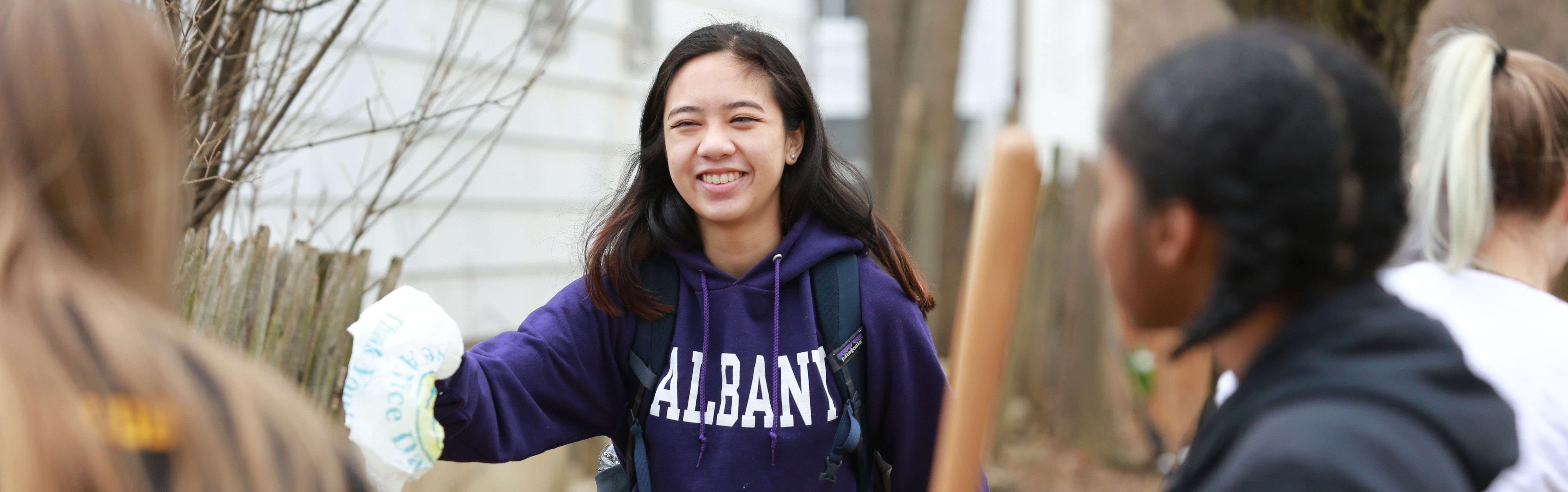 A smiling student wearing a purple UAlbany sweatshirt reaches out to throw away a piece of trash in Albany's Pine Hills neighborhood.