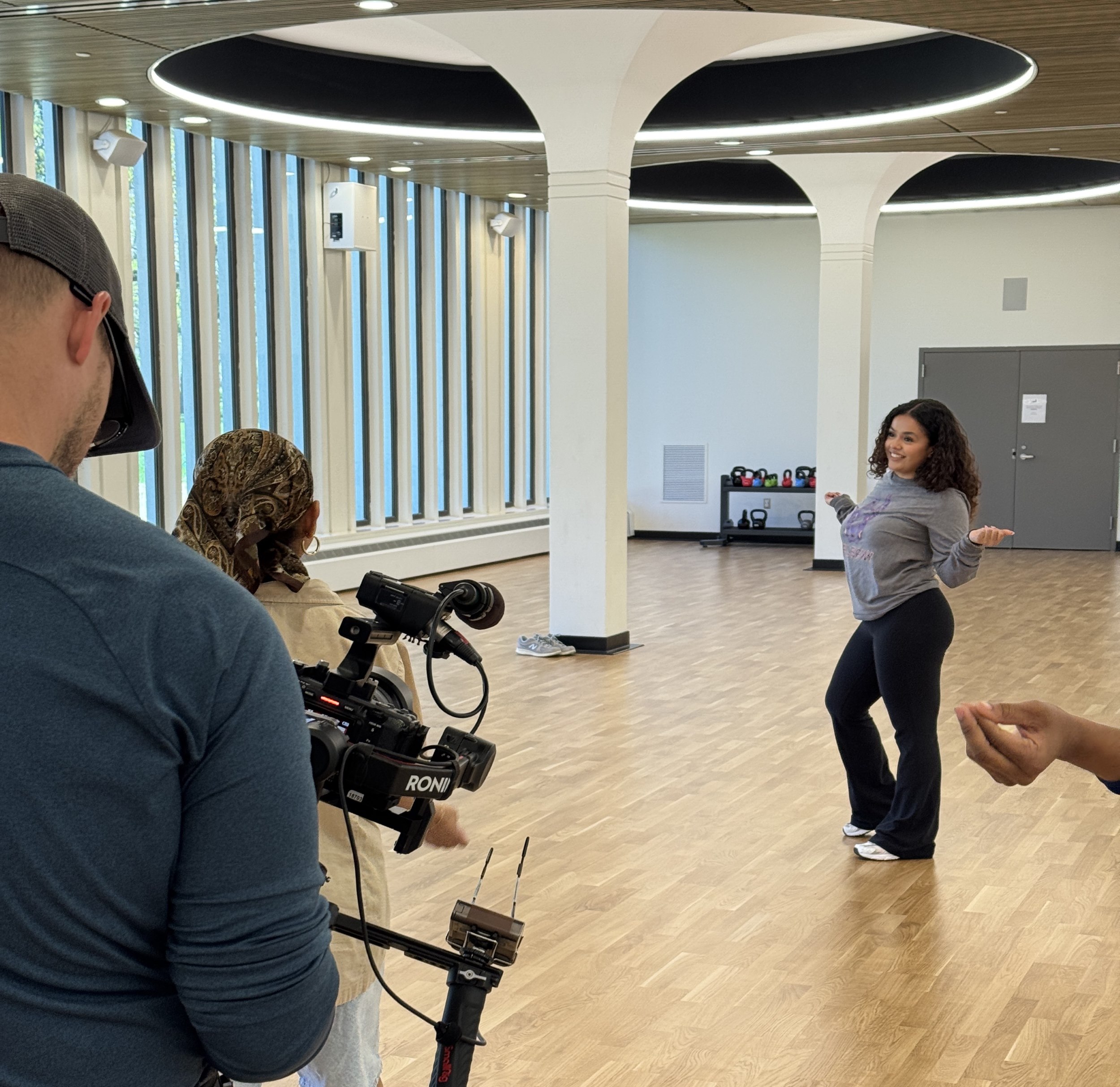 A girl poses in a dance move in a dance studio with her hands raised and at her sides. she wears a gray shirt with the letters UALBANY on it and black leggings, as she is filmed by a man with a camera wearing a portable rigging.