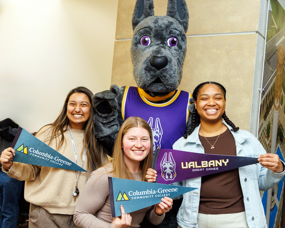 UAlbany mascot Damien the Great Dane with three students holding Columbia-Greene Community College and UAlbany pennants.
