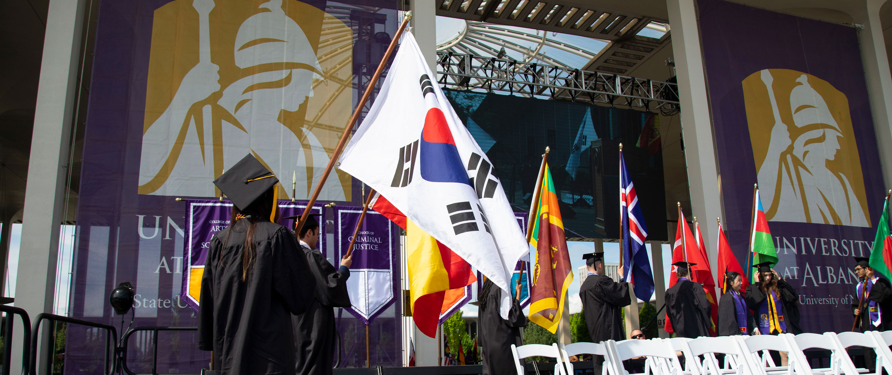 Students in caps and gowns walk onstage carrying flags from their home countries during a practice session for UAlbany's Commencement ceremony.