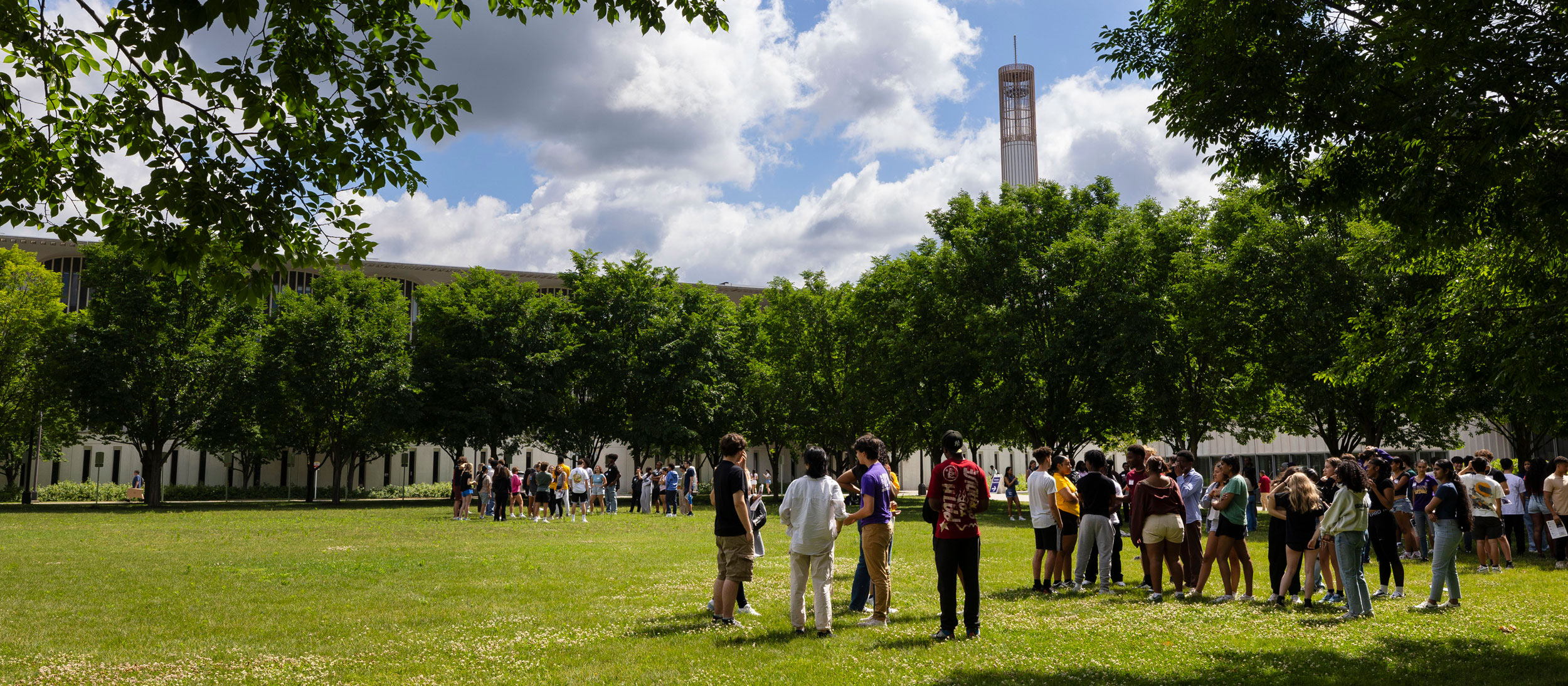 Dozens of students gather outside during a summer Connection Day. The carillon is visible over green tree tops.