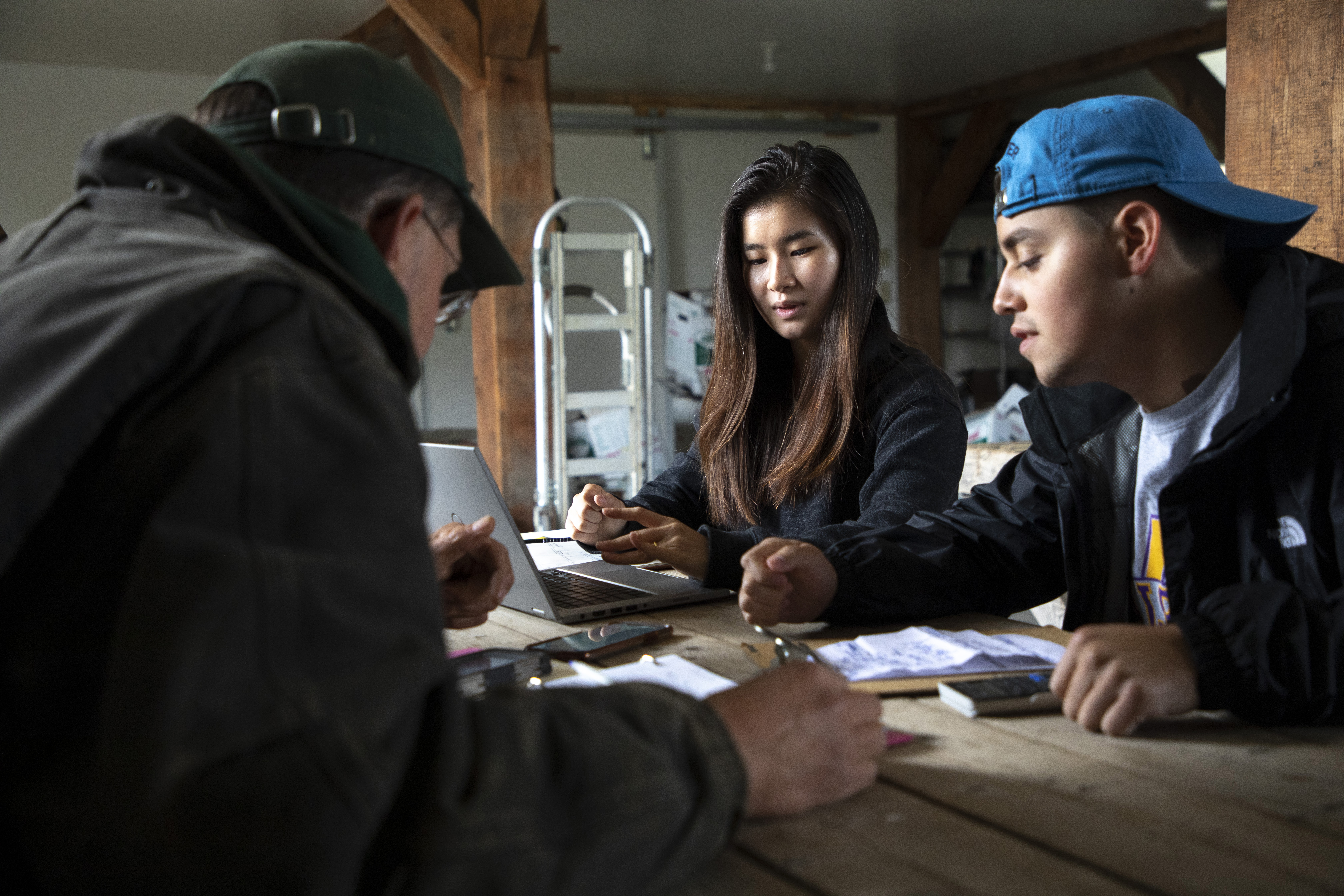 Two students sit inside a barn at a table with the farm owner reviewing notes.