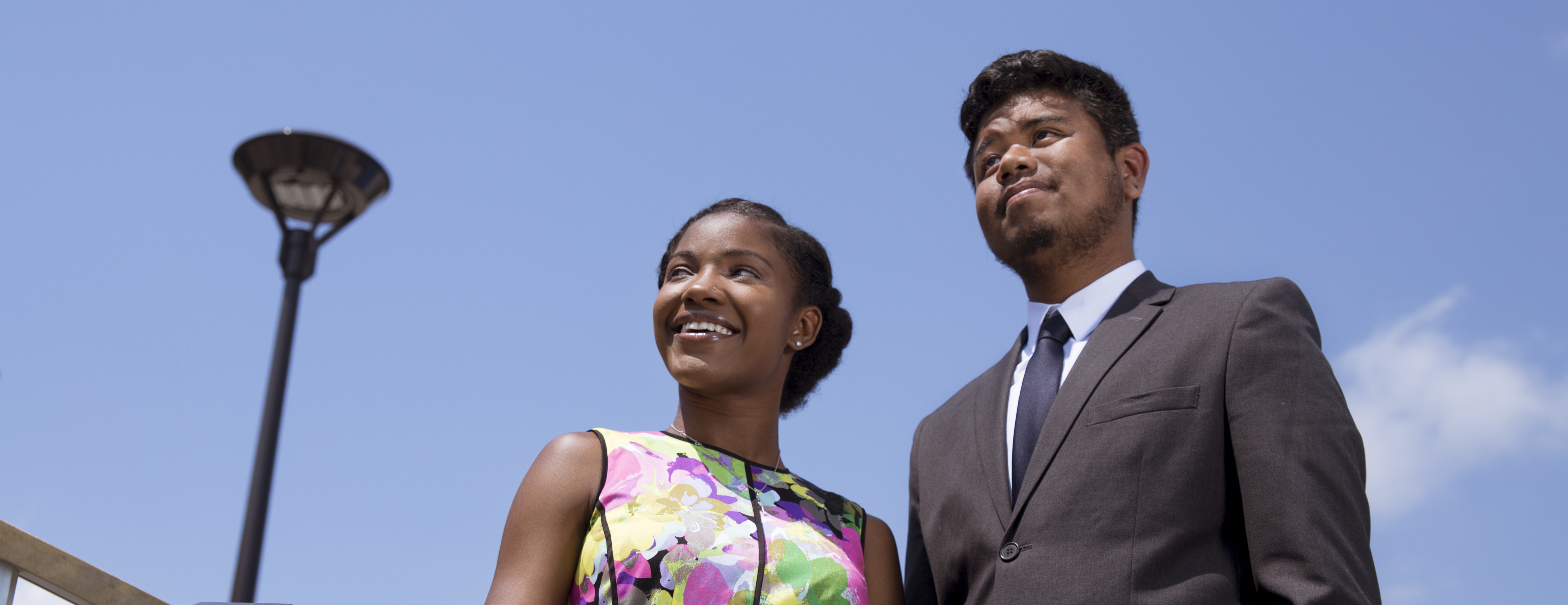 A student wearing a multicolor dress and a student wearing a grey suit smile as they look at a blue sky