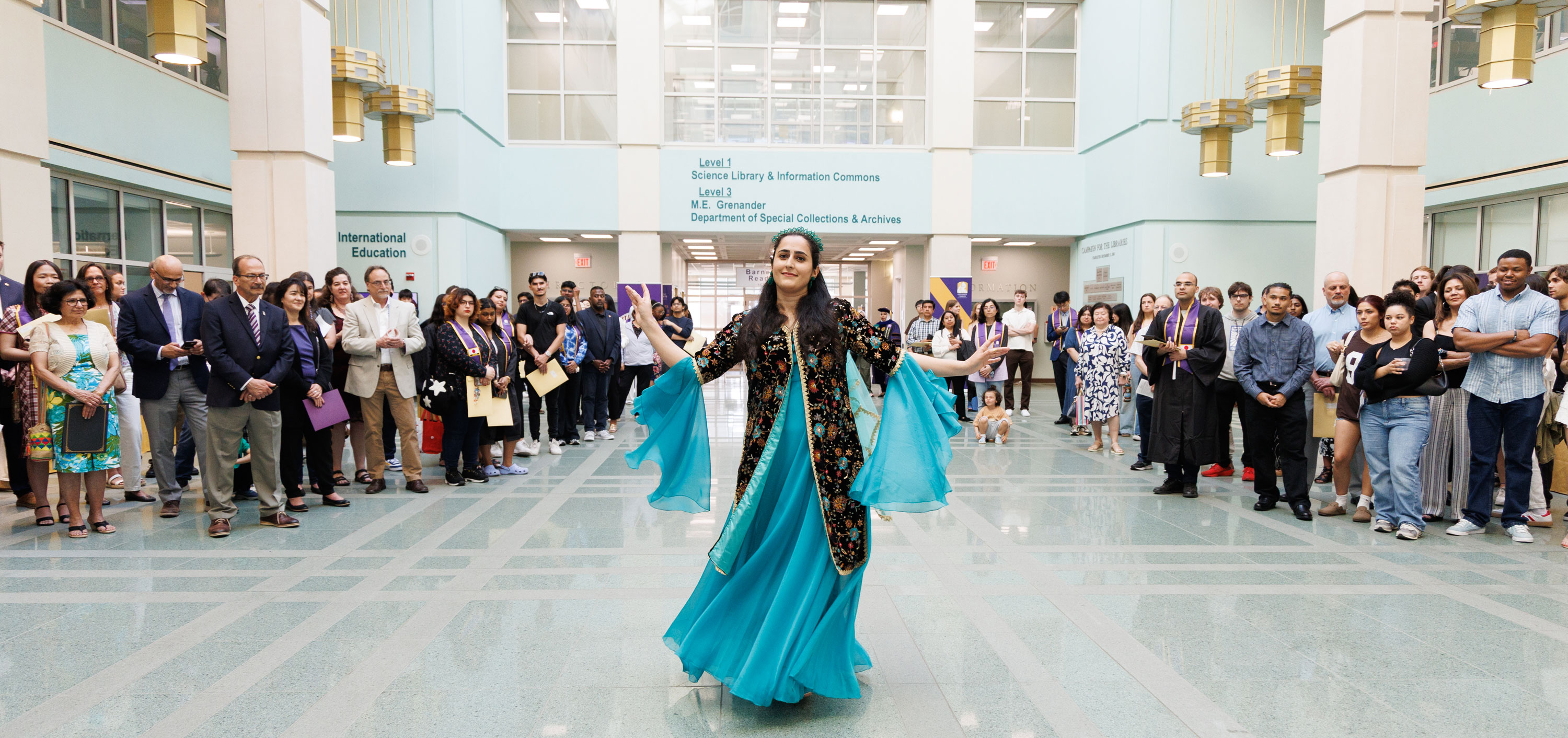 A woman wearing a black and teal dress dances inside the atrium of the Science Library while a gathered crowd watches.