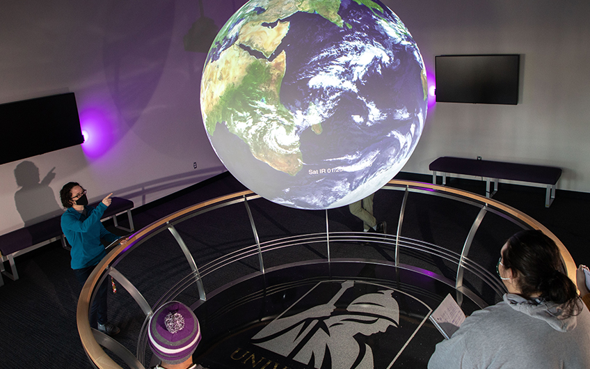 Four people observe a sphere displaying a satellite projection of the earth in UAlbany's Science-On-A-Sphere room