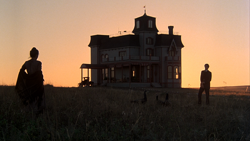 Still from Terrence Malick’s film Days of Heaven (1978), includes house and landscape in shadows with two people standing on either side.