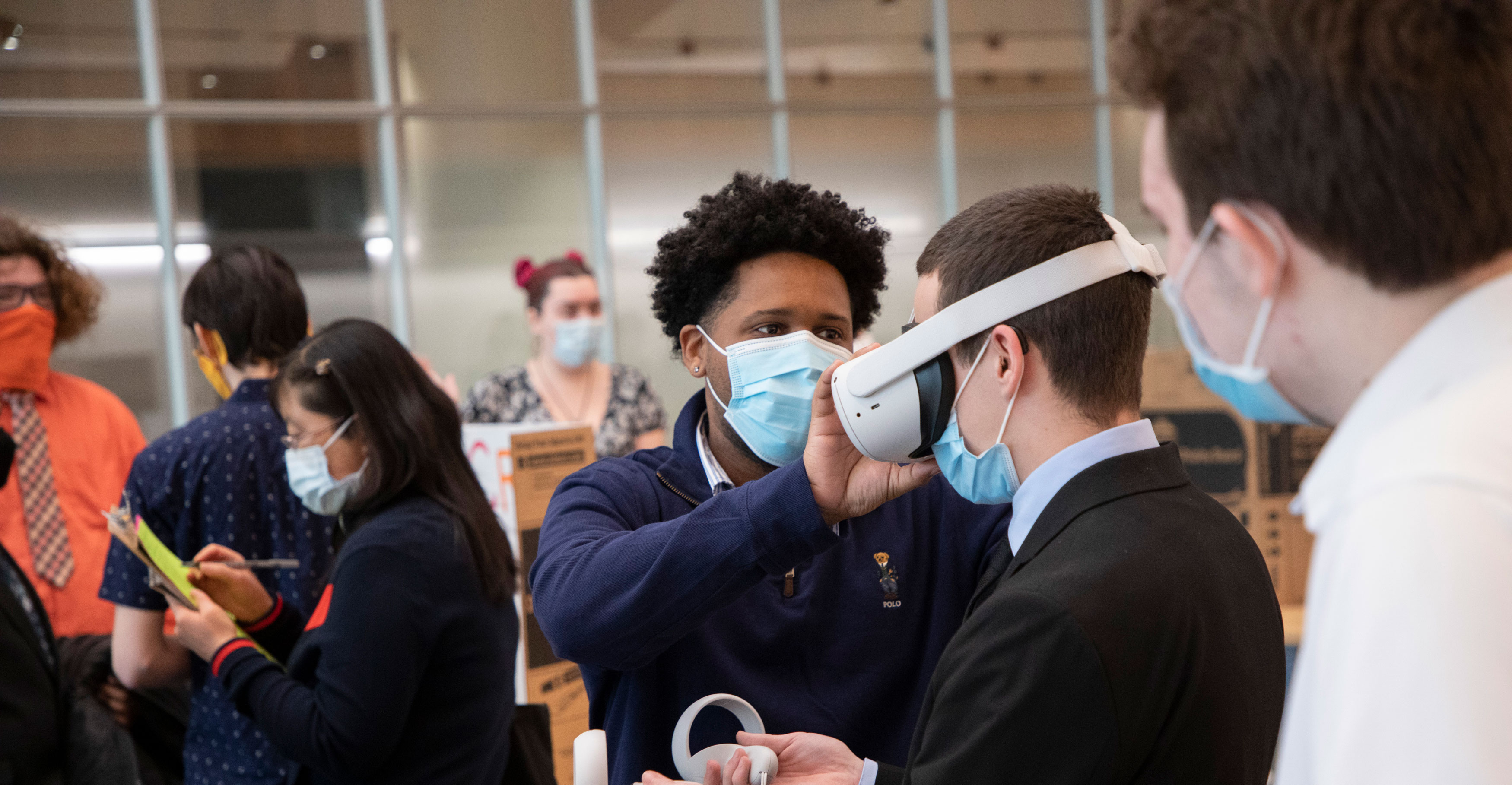 A student helps another person put on a virtual reality head seat during a poster session presentation.