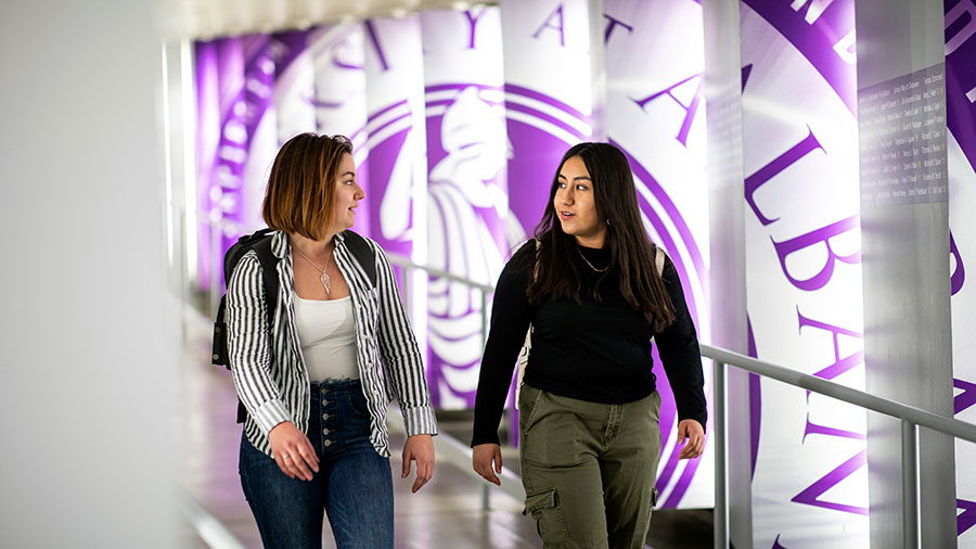 Two students walk through the hallway with the donor wall leading to the Massry School of Business building.