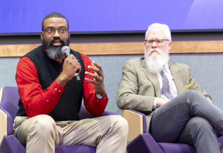 Two bearded men sit on chairs, one speaking into a microphone