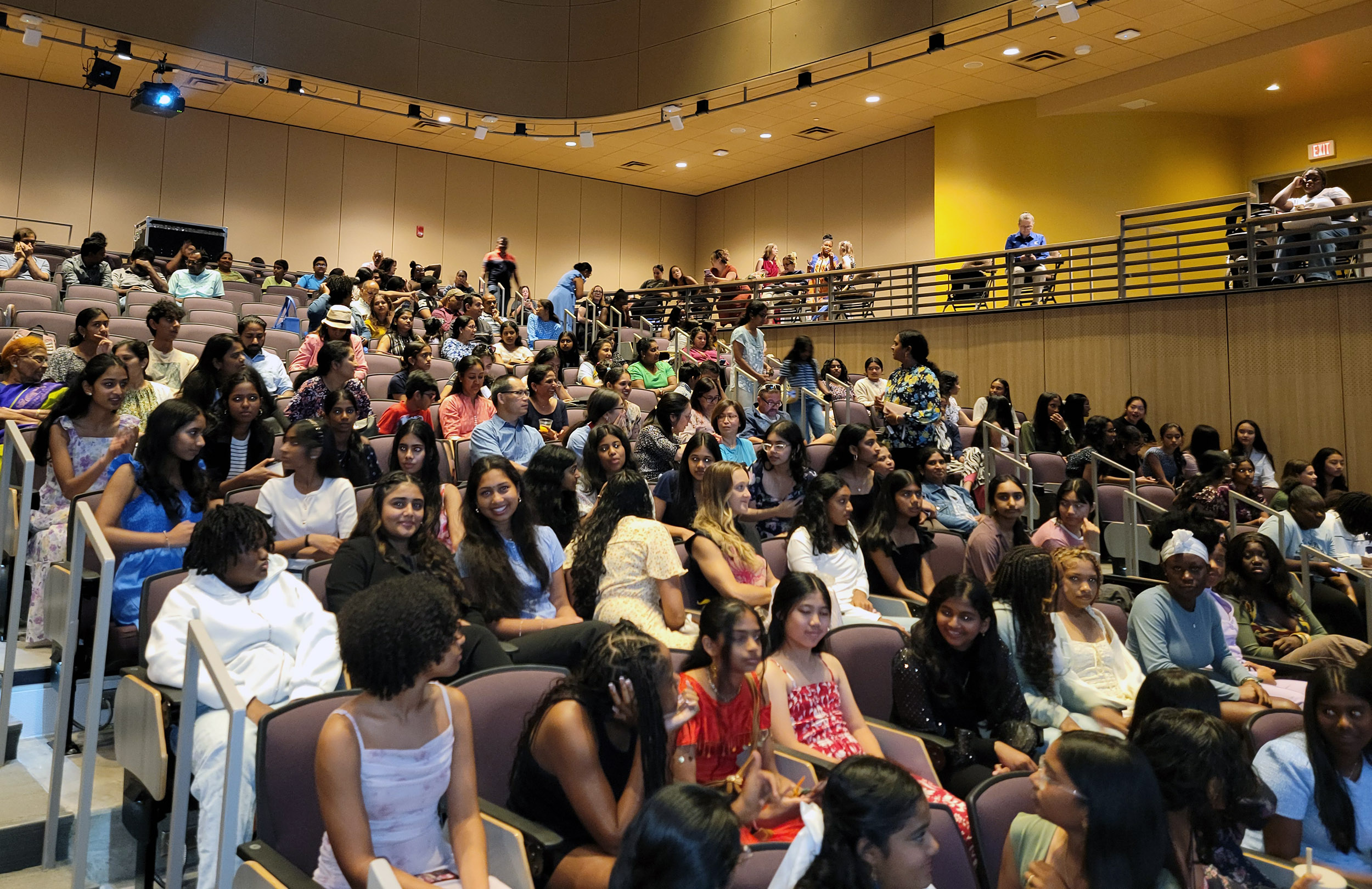 A large gathering of smiling teenage girls and their parents sit in raised seating in an auditorium.