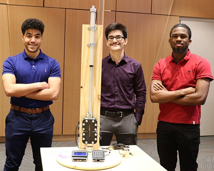 three students stand behind a large wooden and metallic engineering project indoors.