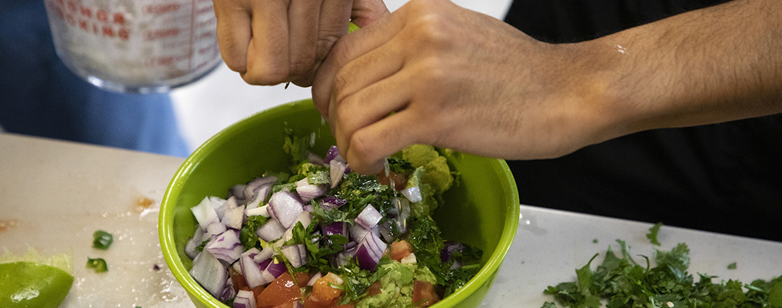 Two hands squeeze a lime into a green ceramic bowl holding unmixed guacamole ingredients.