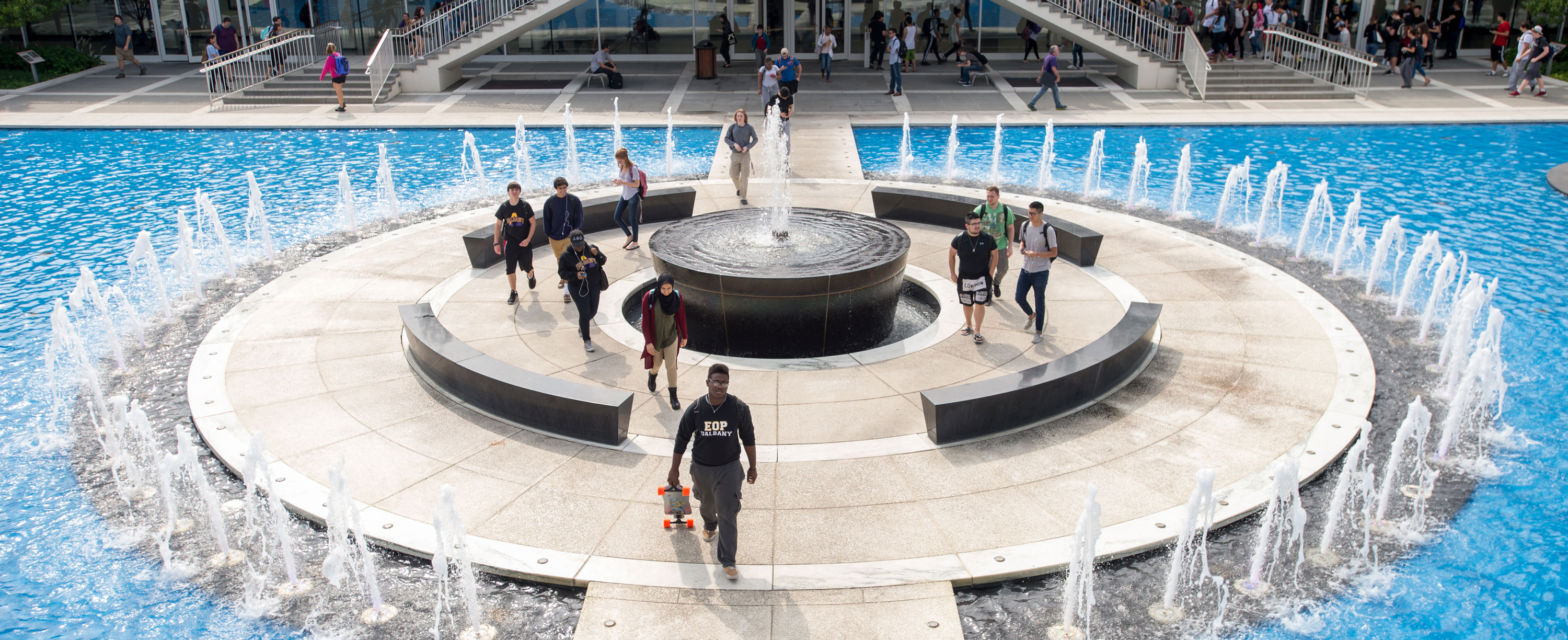 Several students walk along the bridge spanning the Podium Fountain. One student carries a skateboard and wears an EOP shirt.