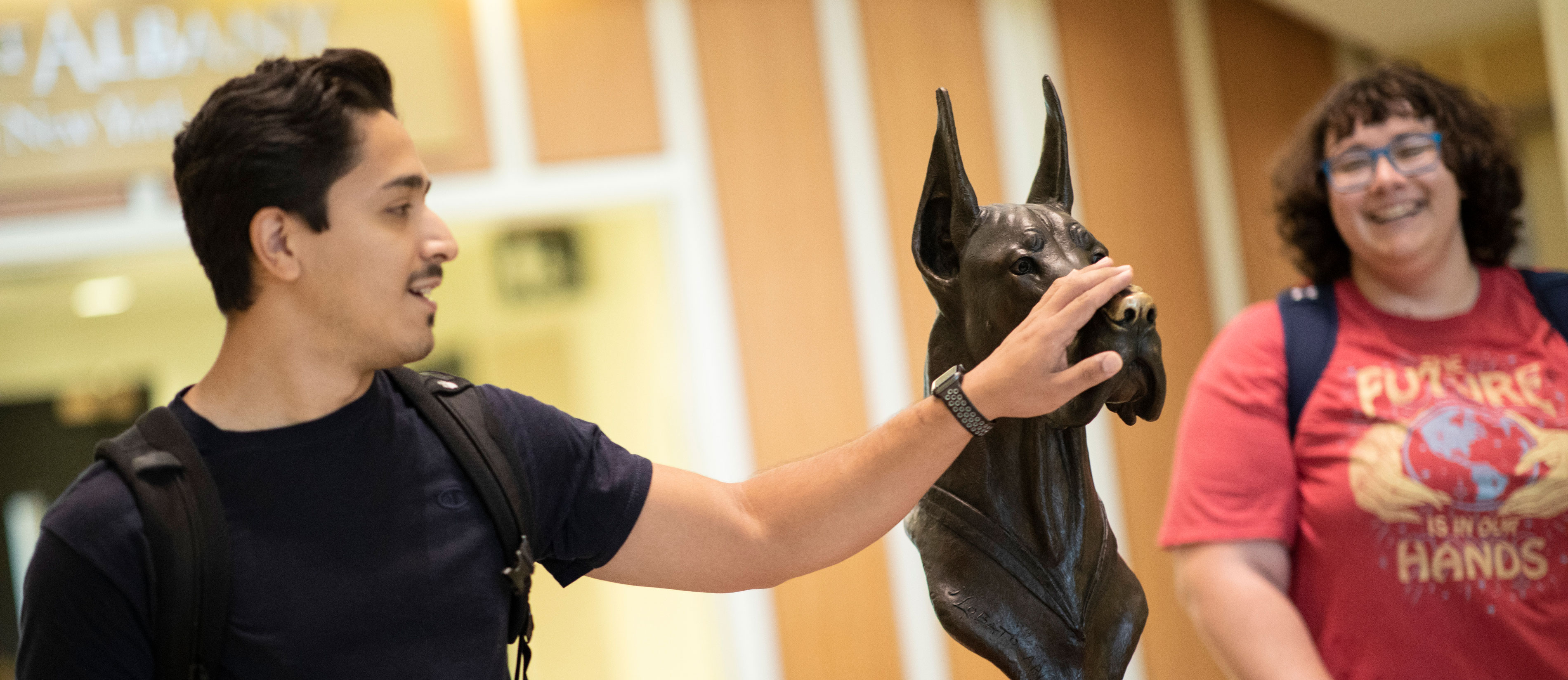 A student reaches out to touch the nose of a bronze Damien the Great Dane statue while another student looks on and smiles.