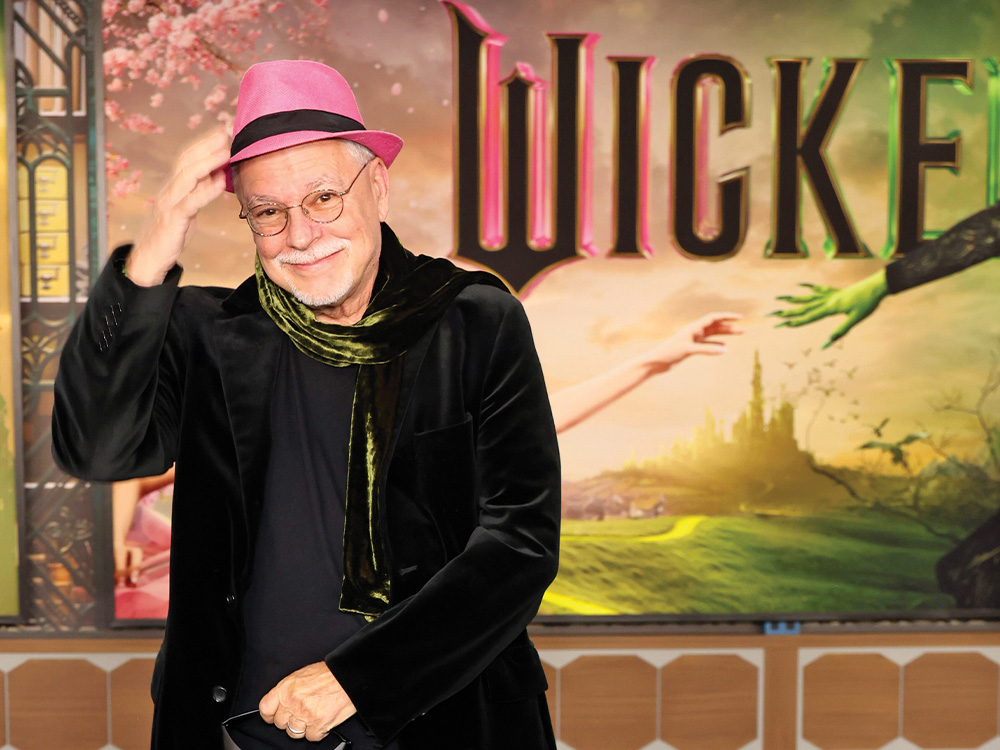 Gregory Maguire with a pink fedora, black jacket and a green scarf standing in front of a backdrop that says wicked with an illustration of oz