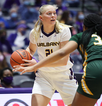 A woman holds a basketball in her hand preparing to dribble against a defender. Her shirt says UAlbany.