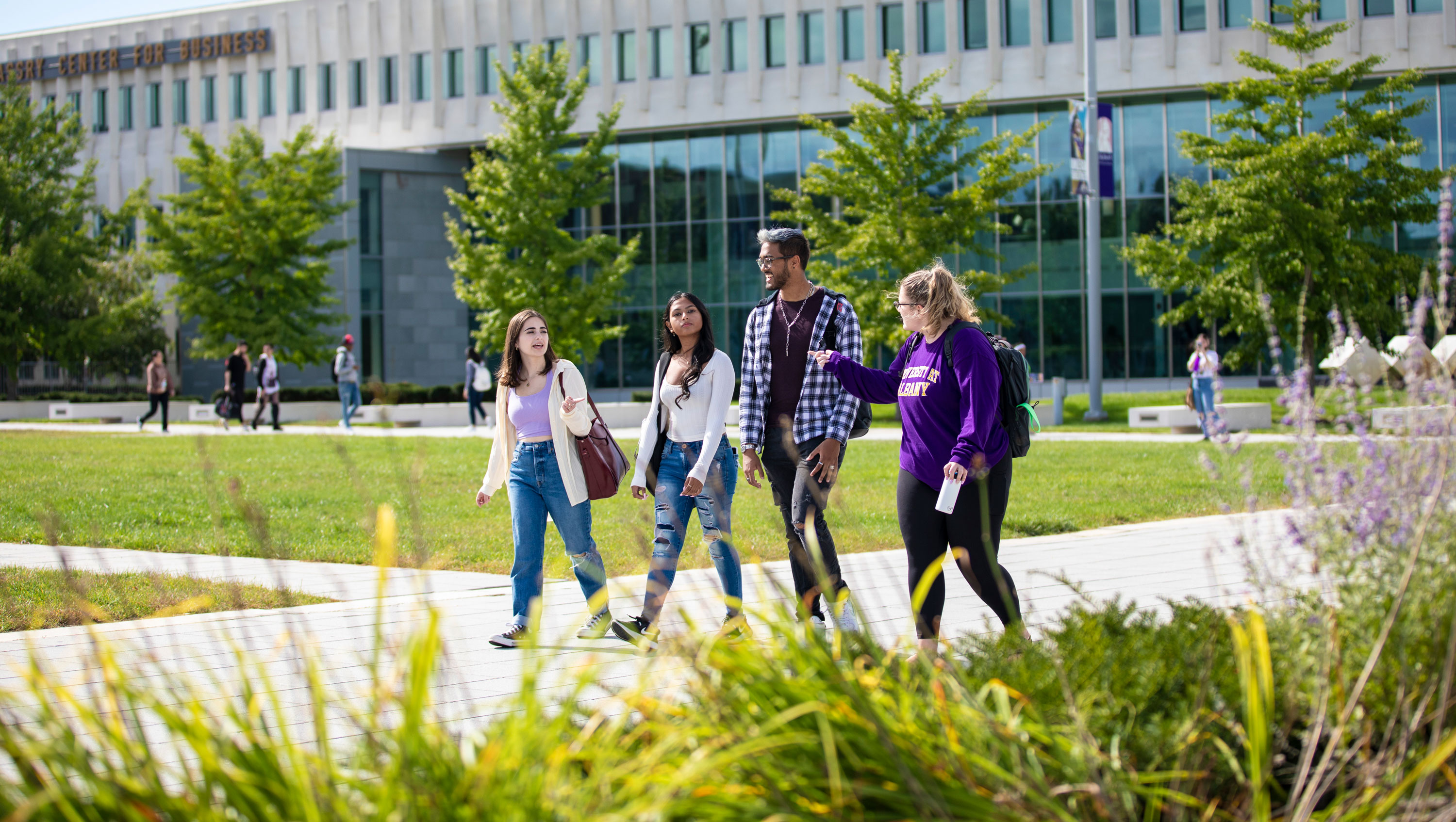 Four smiling students with backpacks walk along a campus pathway on a sunny day.