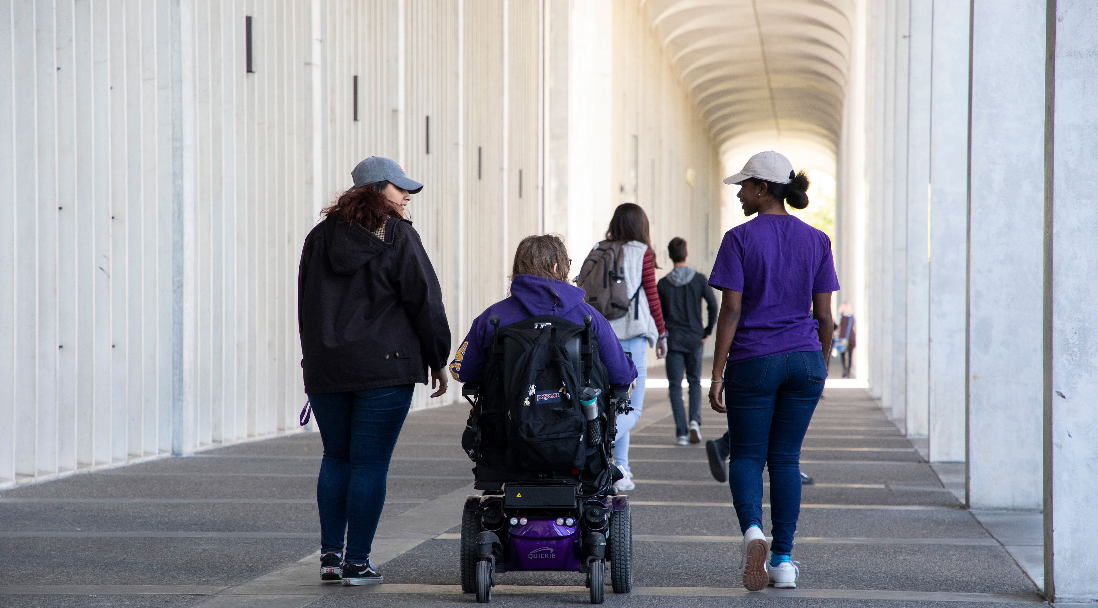 Two students walk on either side of a third student using a motorized wheelchair under the arches of UAlbany's Uptown Campus.