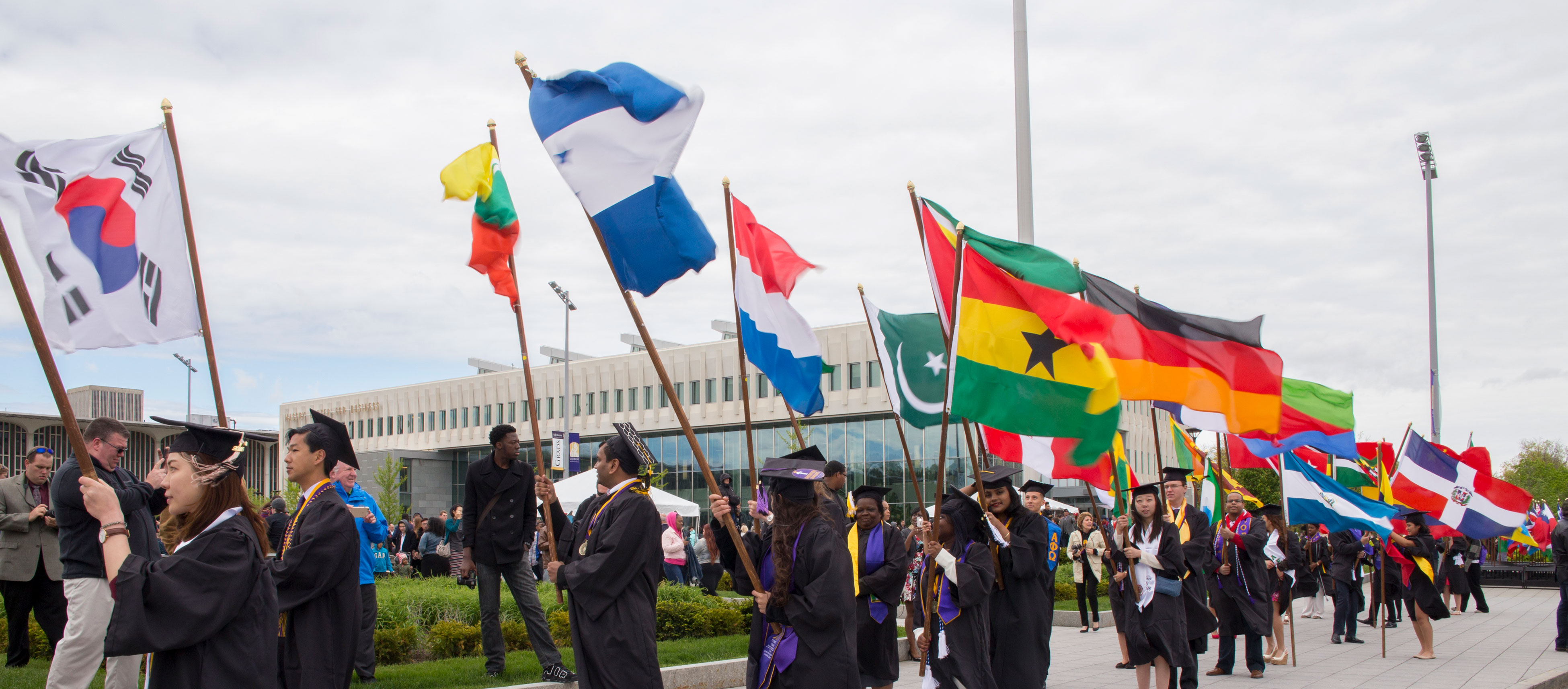 Students wearing graduate caps and gowns walk through campus holding flags from various countries.