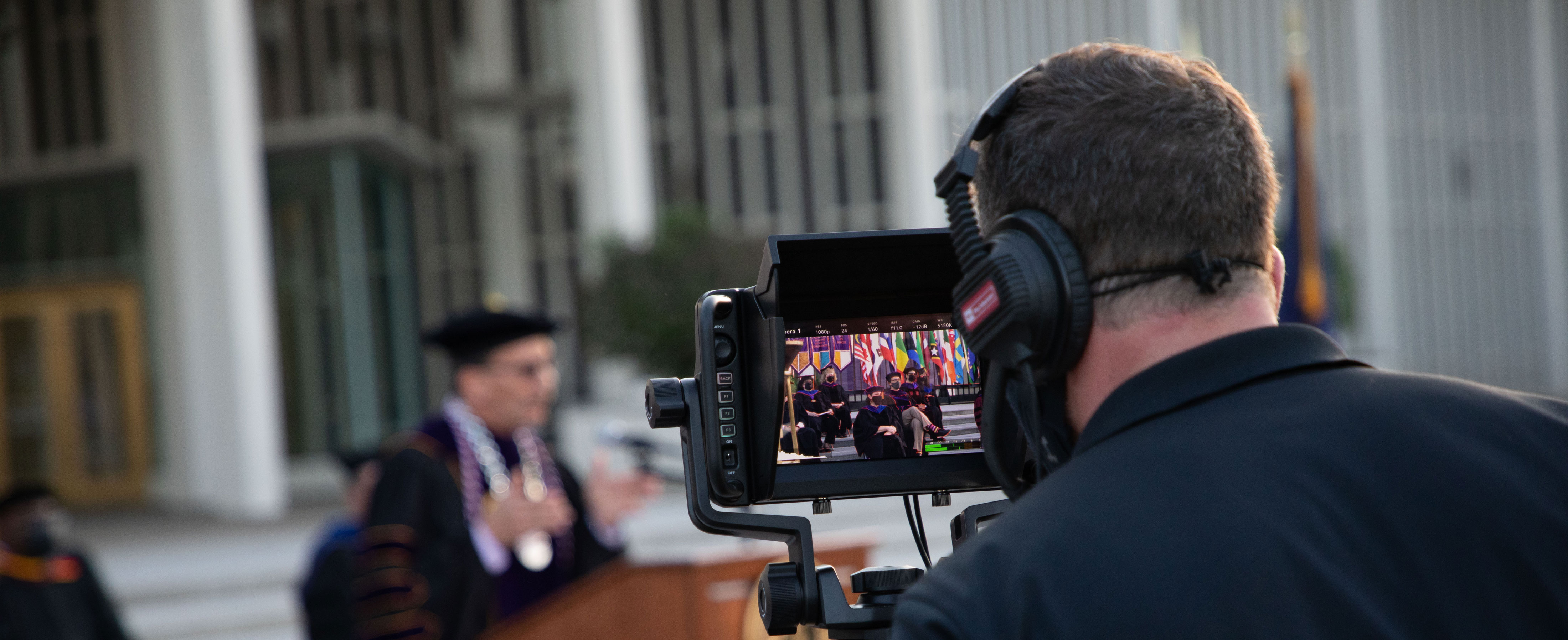 A camera operator wearing headphones looks into a camera display screen as he records Commencement